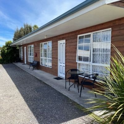 A single-story building with wooden siding, featuring two windows and a white door, represents the charming style found at Castaway Strahan. A small outdoor seating area with two chairs and a table is visible beside the entrance.