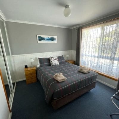 A bedroom at Castaway Strahan with a double bed, neatly made with striped bedding and two folded towels. There are side tables with lamps, a painting on the wall, and a window with sheer curtains.